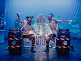 Two men playing drums made of recycled junk on a futuristic stage set.