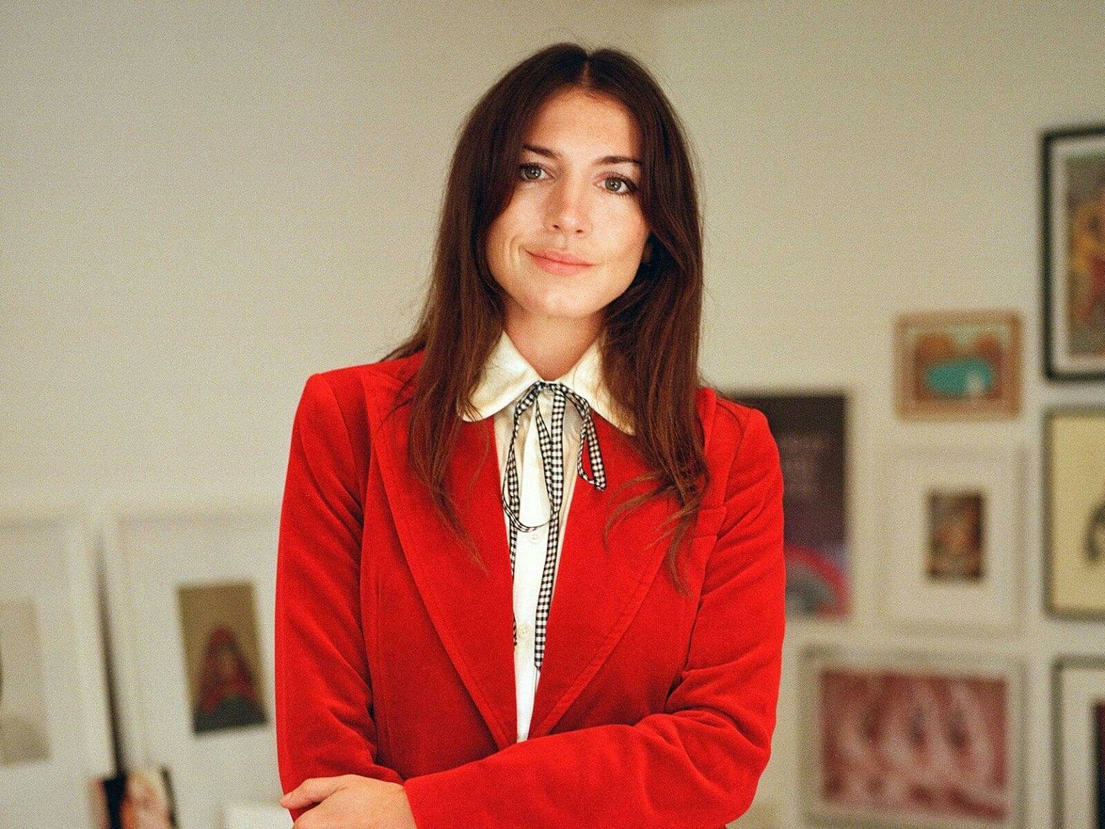 Katy Hessel stands in front of a white wall with several paintings behind her wearing a red suit