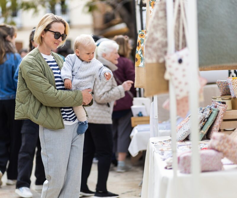 A mother is browsing an outdoor market stall while holding her child in her arms.