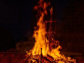 A bonfire burns with flames leaping high on a timber wickerman structure. The background is black