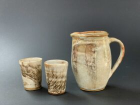 Two cups and a jug are on a grey background. Cream colour glaze with dark brown brushwork decoration