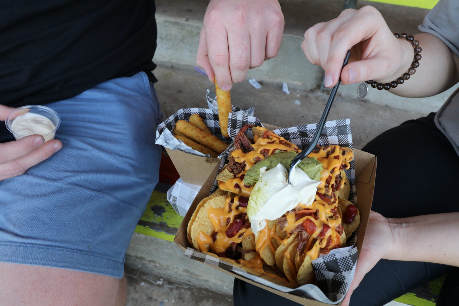 Two hands are shown one picking up a hot chip and one holding a fork poised over a dish of nachos
