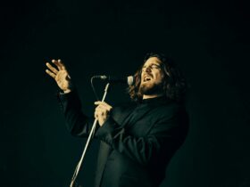 Man with long wavy hair wearing a black shirt singing into a microphone on a stand.