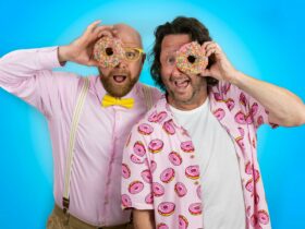 2 male actors in pink shirts against a blue background, looking through a doughnut.