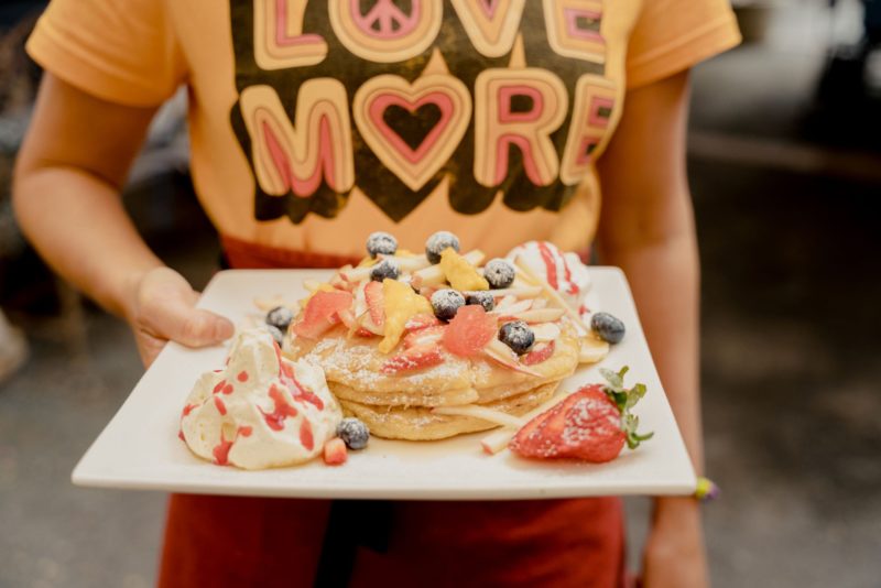 A waitress in a yellow Love More shirt holds a square plate with pancakes and fruit.