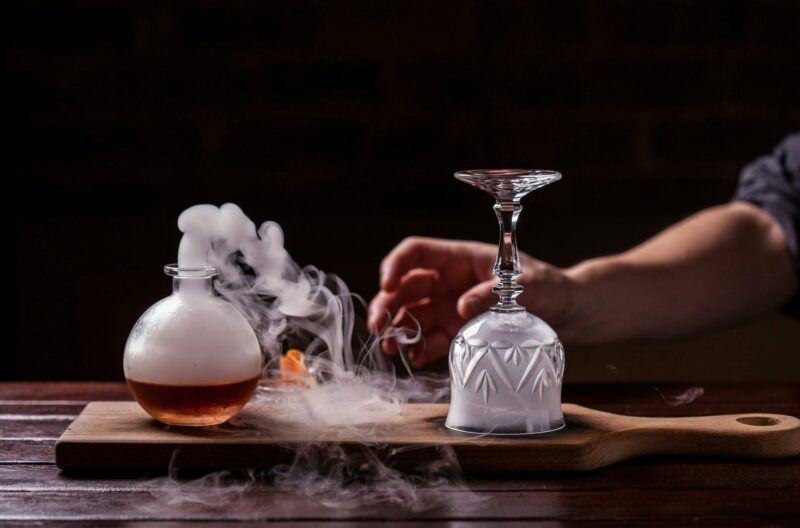A smoking bottle on a wooden board with a hand about to turn over a smoke filled glass
