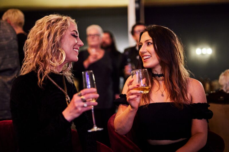 Two lovely ladies enjoy sparkling wine and laugh together in a busy bar.