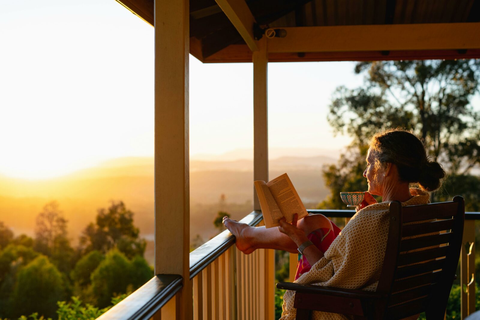a woman reclines on a deck watching the sunrise, drinking tea and reading a book