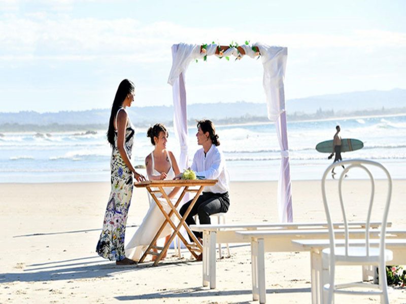 A couple and their celebrant at at a signing table with a wedding arch and ocean in the background