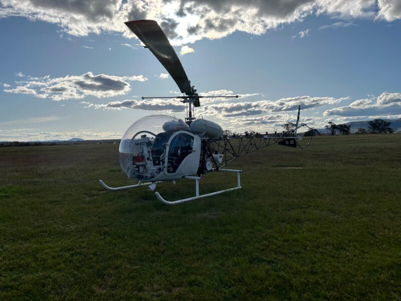 A photo of a Bell 47 helicopter landed in a field, backed by a slightly cloudy blue sky.