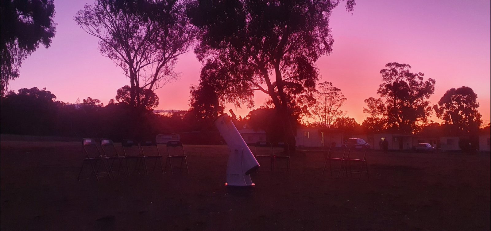 Telescope surrounded by chairs with sunset in the background, through the trees