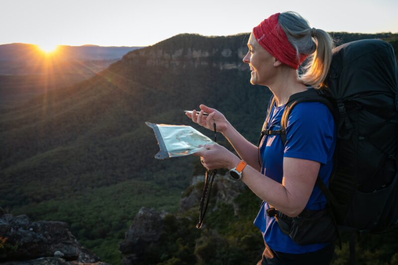 Woman stands on cliff in bush with map and compass looking to horizon