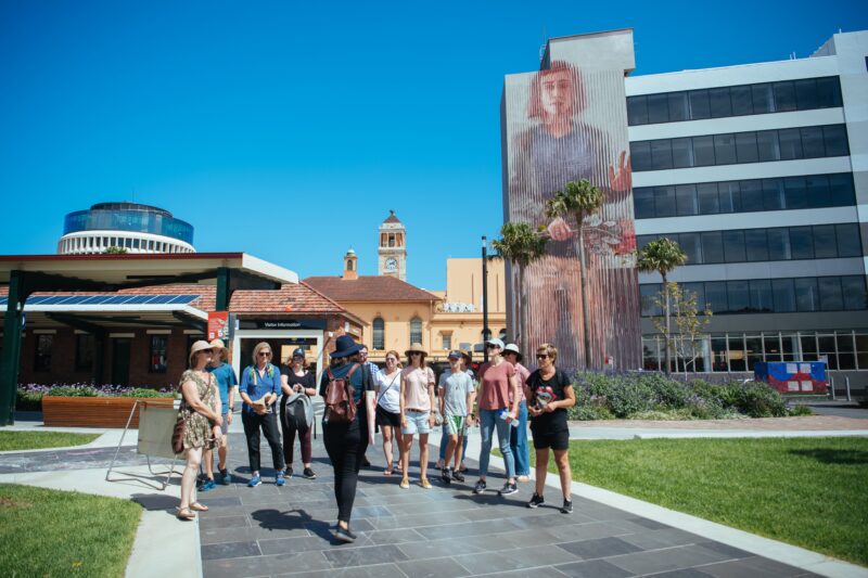 A group of people listen to a tour guide, standing in front of a large mural by Fintan Magee