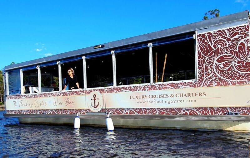 The Floating Oyster Wine Bar. Long pontoon boat side view. Brown patterns with a lady looking out.