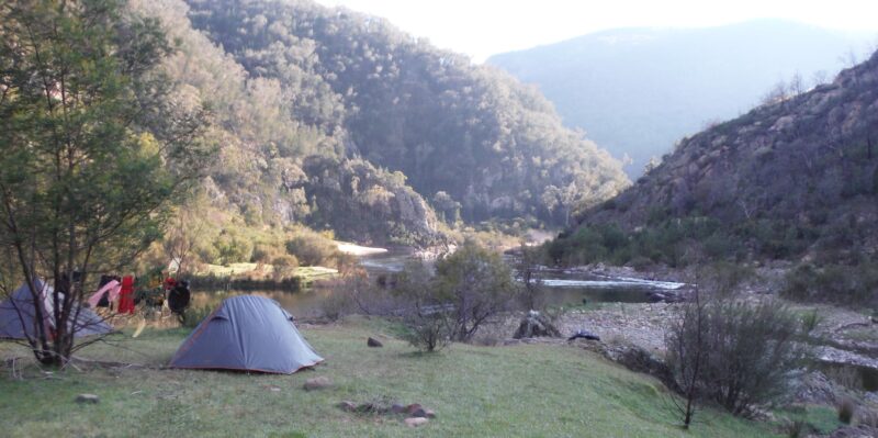 A tent is in the foreground on the edge of the Snowy River, beautiful mountains in the background