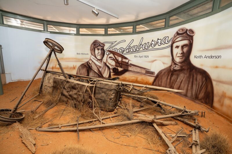 Portraits of Hitchcock and Anderson behind remains of their plane in diorama