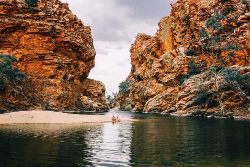 Two friends sitting on the banks of the waterhole with towering red escarpments in the background