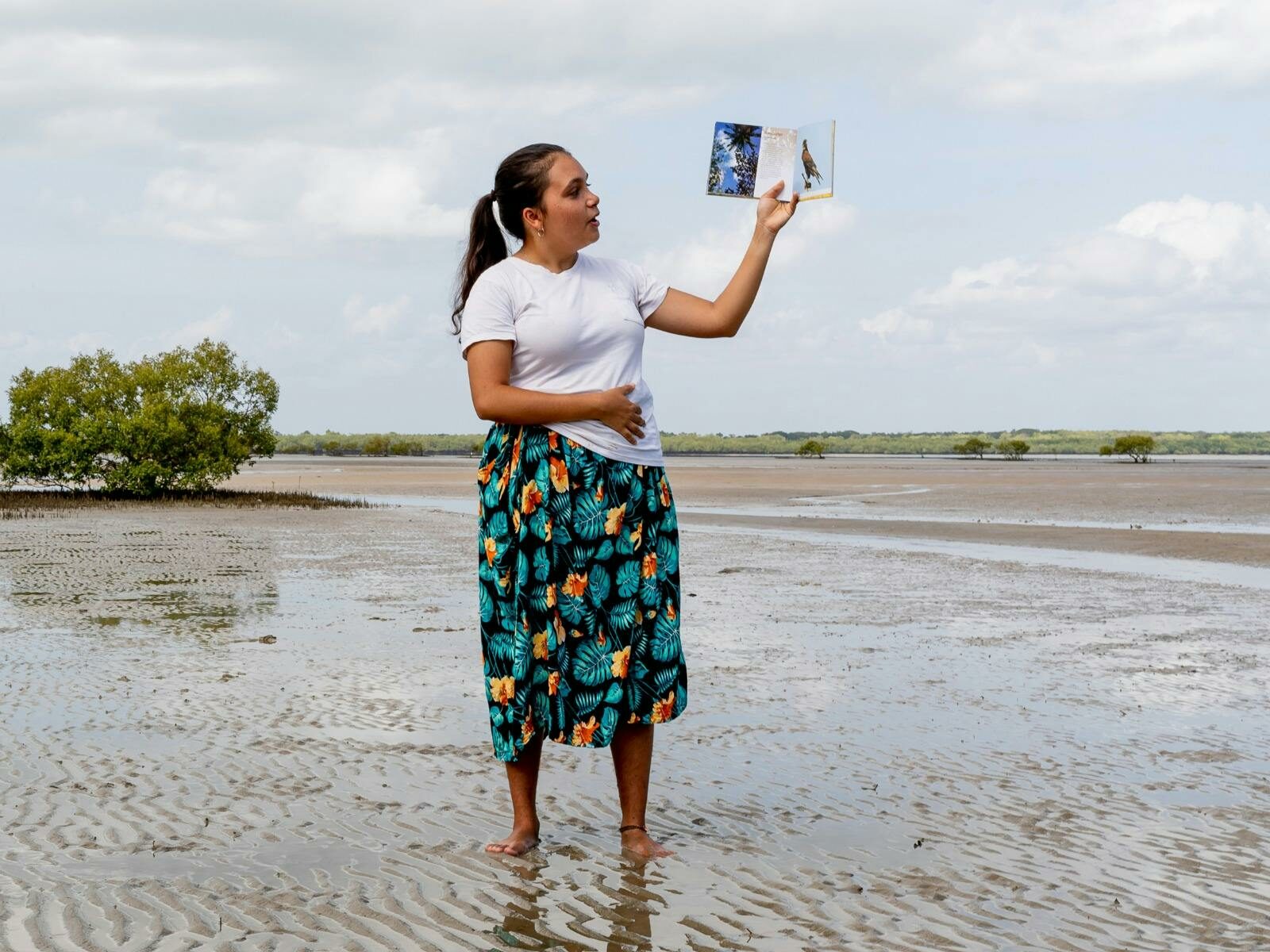 A woman examines a photo while standing on a muddy shore with rippled patterns, wearing a white top
