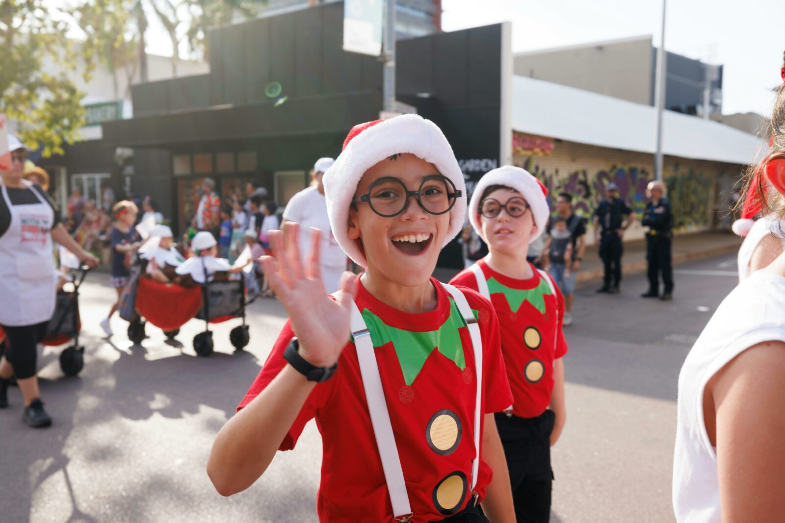 Two children dressed as elves happily wave at the camera, spreading holiday cheer.