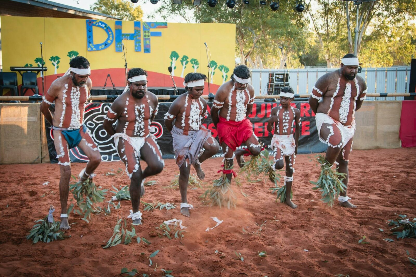 5 Indigenous men & 1 child dressed in traditional clothing & body paint dance in front of a stage.