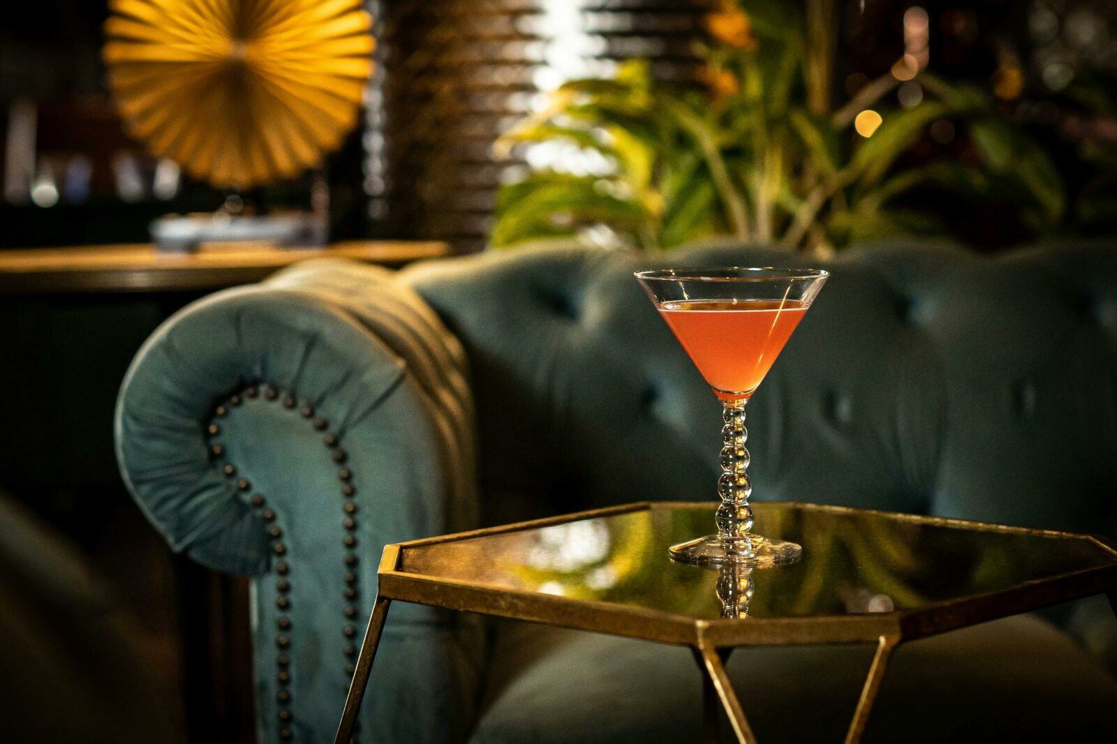 A velvet green couch as the backdrop of a cocktail on a glass table inside the cocktail lounge