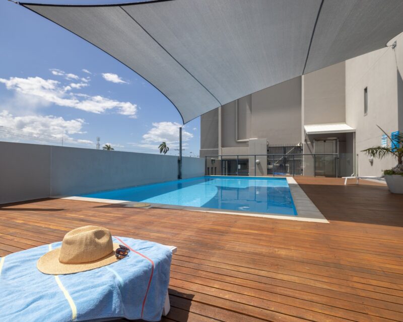 Swimming pool with wooden deck surrounding two sides. Towel, hat and sunglasses on a pool lounge.