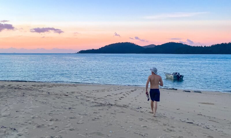 A man walking on a beach towards small boat in water and sunset over island hills in the distance