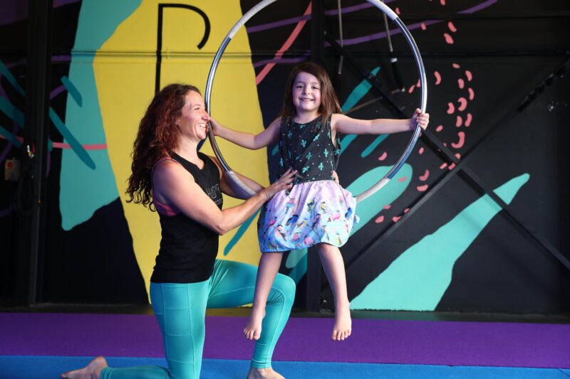 Kids Aerial Classes - Teacher is supporting a young girl who is sitting on an Aerial hoop.