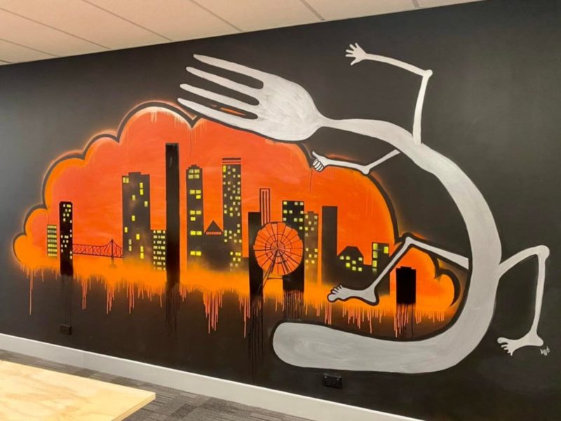 Mural on a black wall of the skyline of Brisbane being cuddled by a large fork with arms and legs