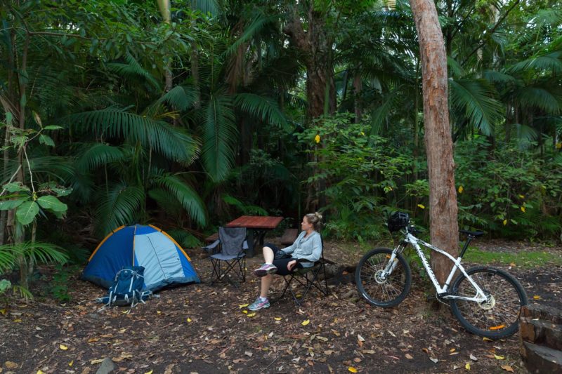 Person sits near her small tent and mountain bike surrounded by tall forest.