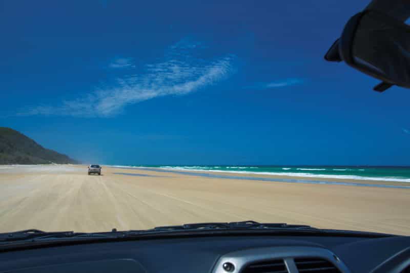 View through car window of driving along sandy beach towards headland, Cooloola.
