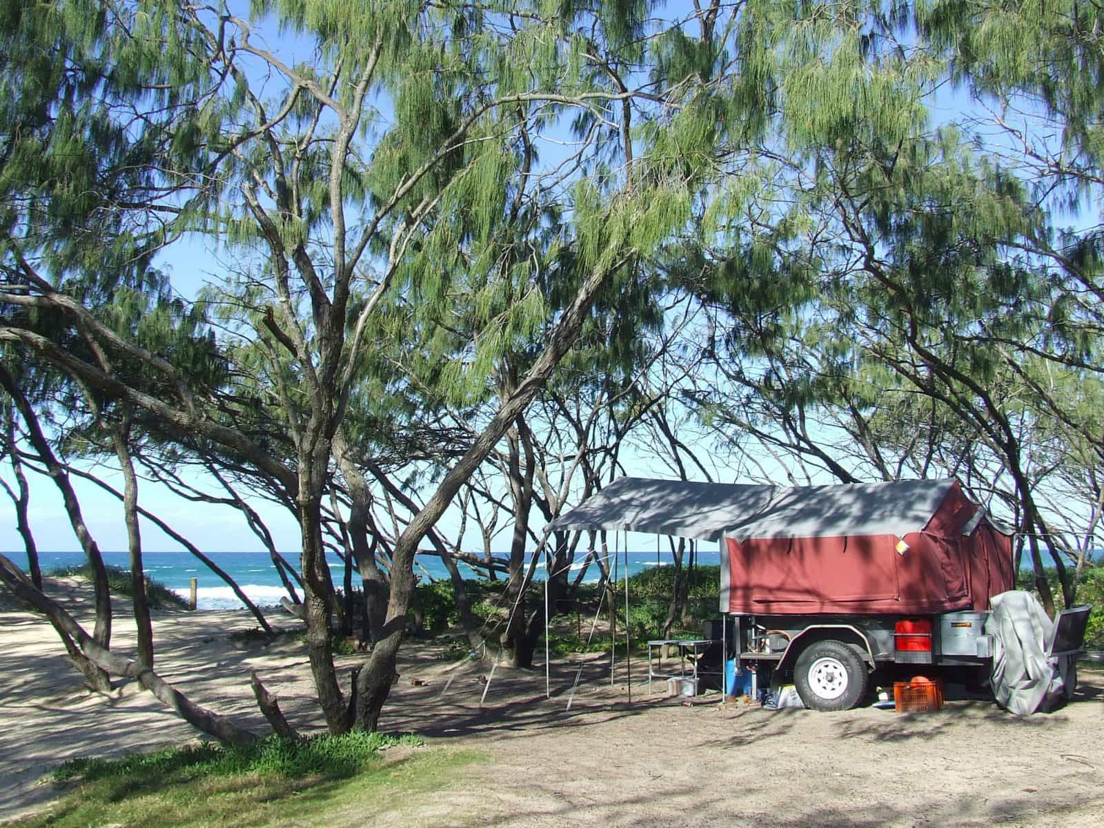 A red camper trailer is parked amongst the casuarinas behind the beach,