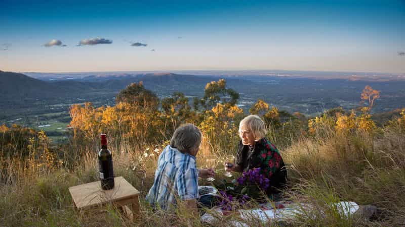 Older couple sitting on a grass hill with wine overlooking valley at dusk