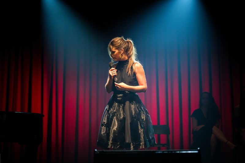 Female in black dress on stage with head to the side, holding microphone, in front of red curtians.
