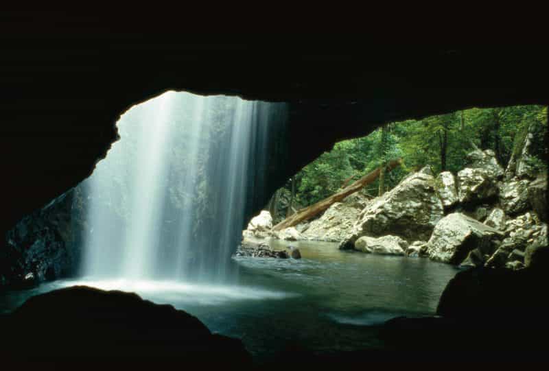 A sheet of white water, backlit by sunlight, falls into a dark rockpool, underneath a rock arch.