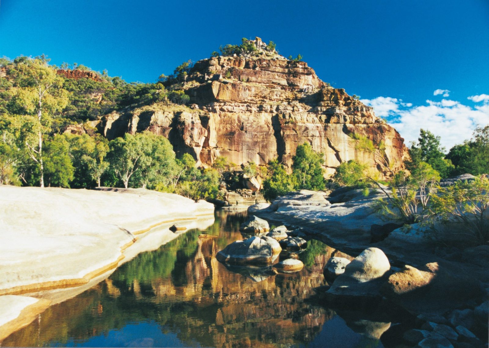 A pyramid-like sandstone formation towers over a small creek with boulders, fringed by vegetation.