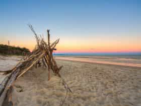 Large triangular tent made out of sticks and branches on the beach at sunset