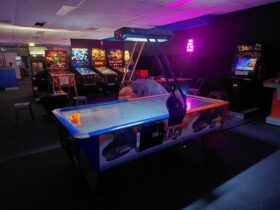 A lit up air hockey table in a dim lit room, pinball machines, arcade machines and a neon sign