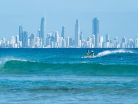 Two women surfing in blue ocean with city skyline in background.