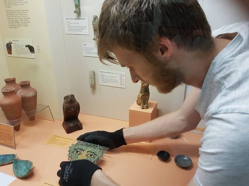 A view into the Egyptian Display Case showing staff with gloves arranging artefacts and labels .