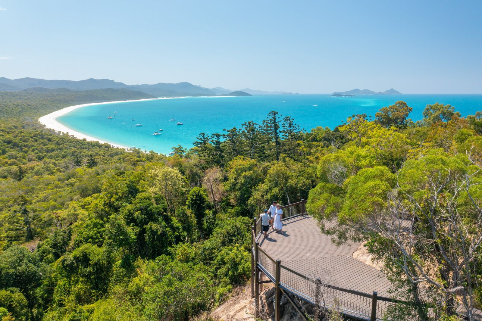 People on a hilltop lookout point overlooking the trees, long stretch of white sandy beach and ocean