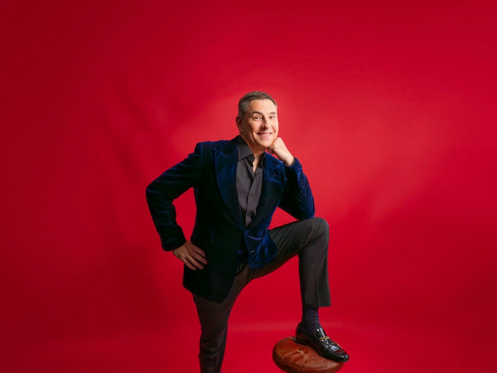 David Walliams behind a red background with his leg on a stool leaning on his knee smiling in a suit