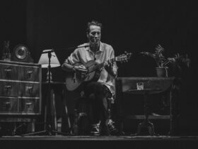 Marlon Williams - person sitting in the middle of a stage, holding a guitar surrounded by furniture