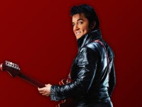 Elvis Presley impersonator standing in a leather jacket holding a guitar in front of a red wall