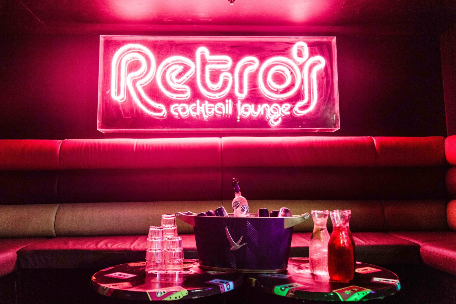 A lounge at a bar with an ice bucket of drinks and alcohol with a neon light behind that says Retros