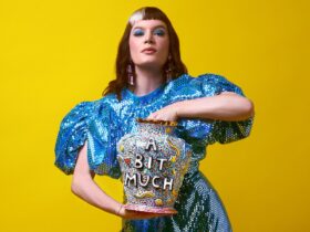 A person in a shiny blue dress holds a large, colourfully adorned vase that says "A BIT MUCH" on it.