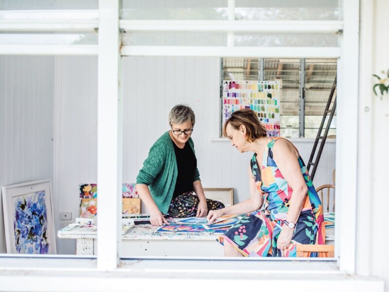 Two women sitting on a table looking at artwork viewed through a window