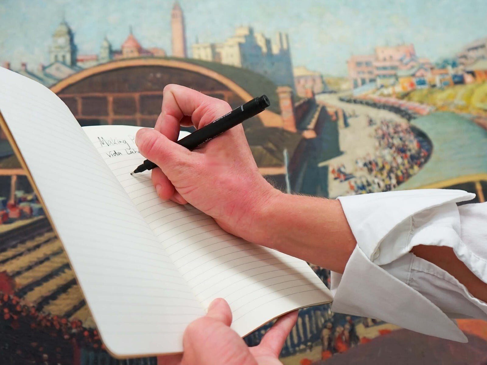 A hand holding a black pen writing in a notebook, a painting in the background.