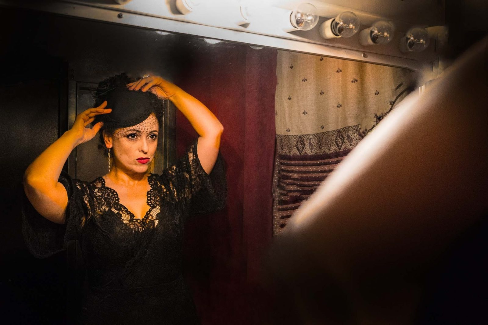 A person wearing red lipstick, a race day hat and black lace edged dress, stares into a mirror
