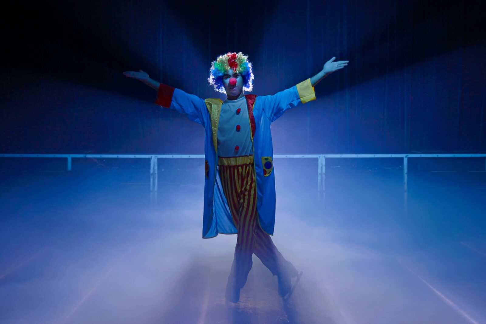 1 clown standing in the middle of the ice skating rink under a spotlight with his arms open wide
